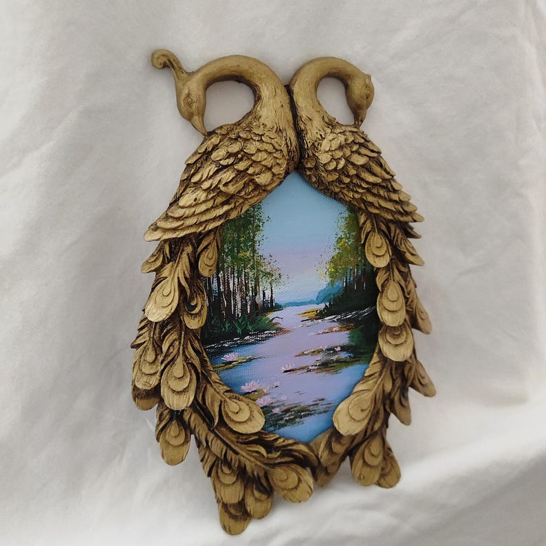 May include: A decorative oval-shaped wall art piece featuring a landscape painting framed by two gold-colored peacock figures. The painting depicts a river scene with trees and a blue sky. The frame has detailed feather and bird head carvings.