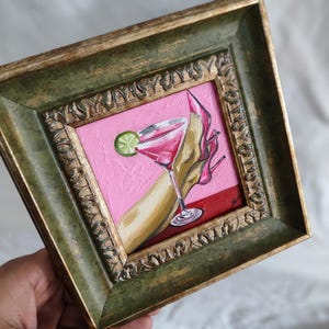 May include: Square framed painting of a pink cocktail with a lime wedge, a high heel, and a hand. The painting has a pink background and a red base. The frame is ornate and green.
