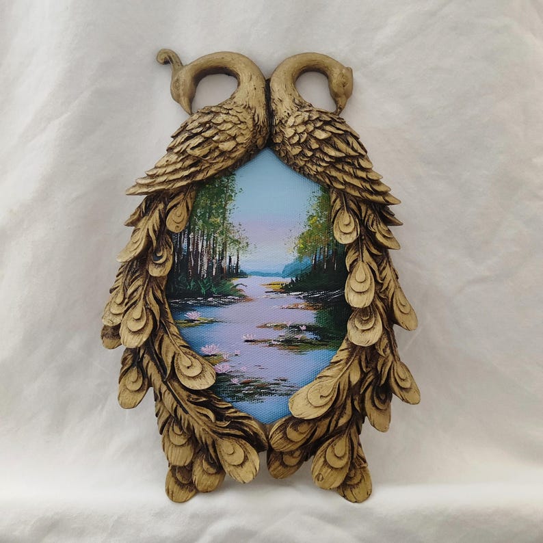 May include: A decorative wall hanging featuring a landscape painting framed by a gold-colored peacock design. The painting depicts a serene river scene with trees and water lilies. The frame is intricately detailed.