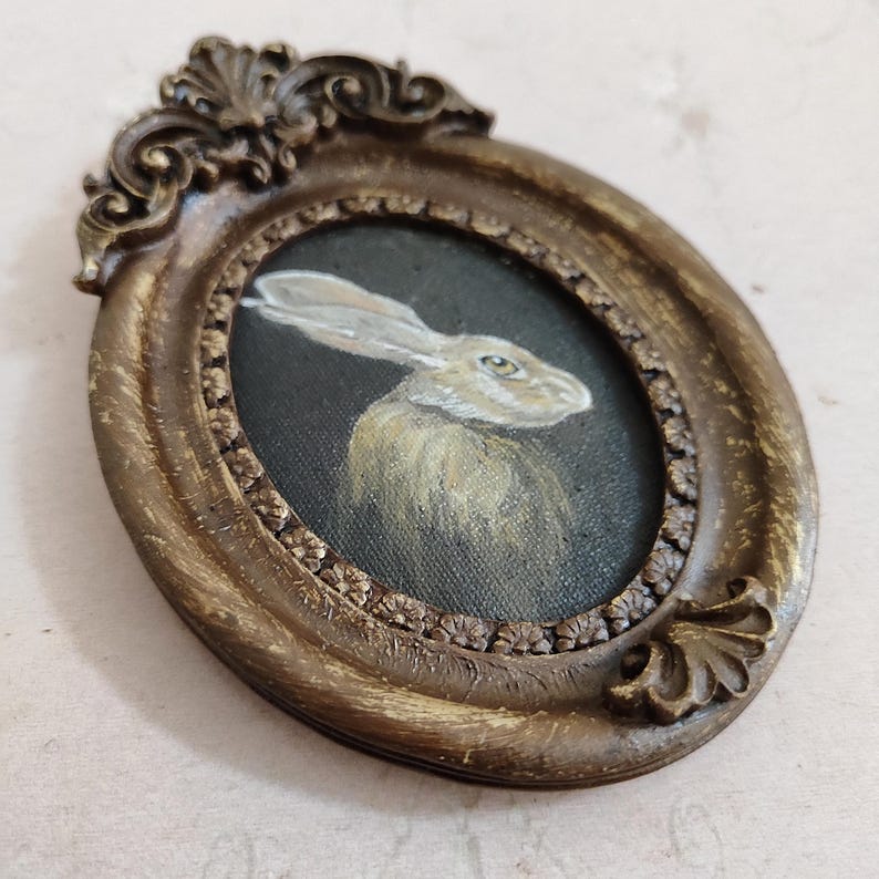 May include: Oval antique-style frame with a painting of a hare. The painting shows a light brown hare against a dark background. The frame is a distressed gold colour with ornate detailing.