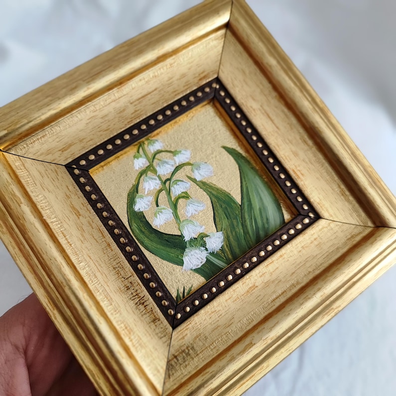 Peut inclure: Petite peinture encadr&eacute;e repr&eacute;sentant des fleurs de muguet. L'&oelig;uvre pr&eacute;sente des fleurs blanches en forme de cloche et des feuilles vertes sur un fond dor&eacute;. Le cadre dor&eacute; a une bordure int&eacute;rieure marron fonc&eacute;. Dimensions: environ 10 cm x 10 cm.