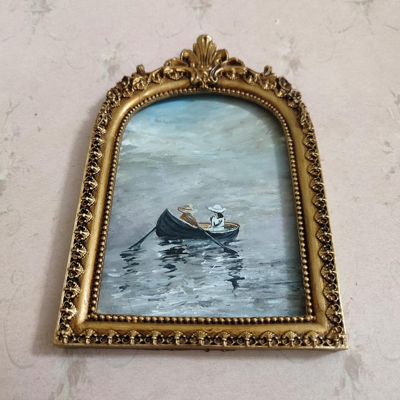 Puede incluir: Una pintura enmarcada en oro de dos personas en un bote de remos en un lago. La pintura es de estilo vintage y tiene una paleta de colores suave y tenue.