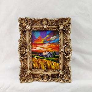 May include: A framed oil painting depicts a vibrant sunset over a rural landscape. The artwork features a house, a field of wheat, and a colorful sky with shades of orange, purple, and blue. The ornate, gold-colored frame adds a classic touch.
