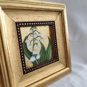 Peut inclure: Peinture encadr&eacute;e de fleurs de muguet. L'&oelig;uvre pr&eacute;sente des fleurs blanches en forme de clochettes avec des feuilles vertes sur un fond dor&eacute;. Le cadre est dor&eacute; avec une bordure int&eacute;rieure marron fonc&eacute;.