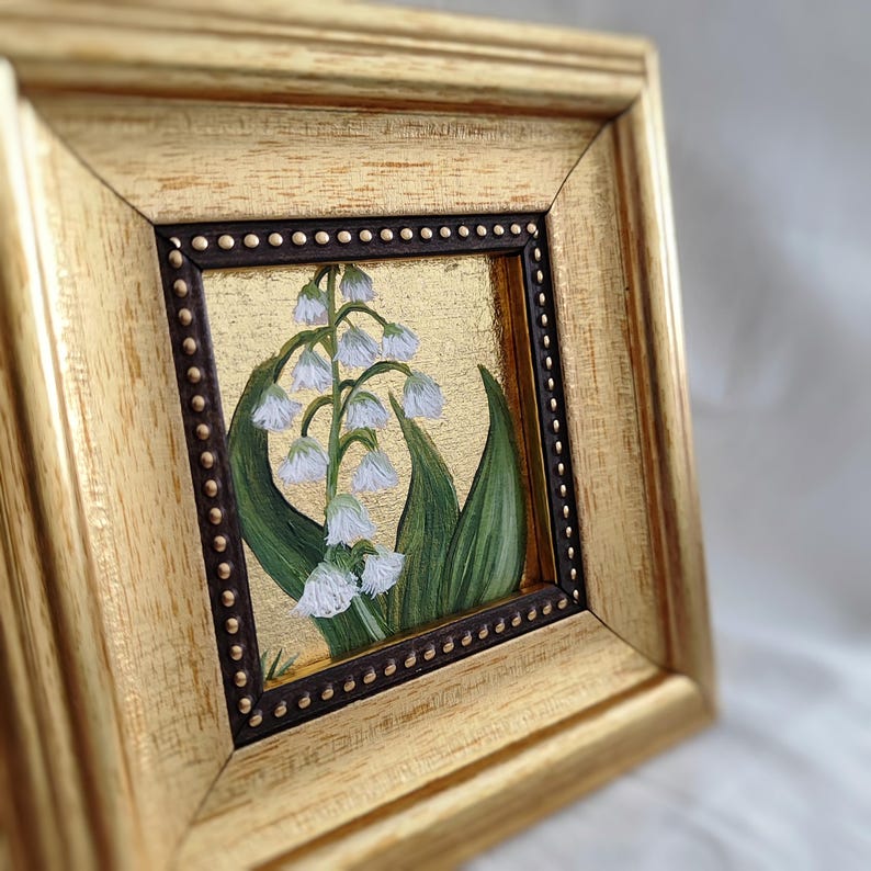 Peut inclure: Peinture encadr&eacute;e de fleurs de muguet. L'&oelig;uvre pr&eacute;sente des fleurs blanches en forme de clochettes avec des feuilles vertes sur un fond dor&eacute;. Le cadre est dor&eacute; avec une bordure int&eacute;rieure marron fonc&eacute;.
