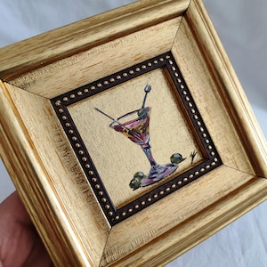 May include: A small painting of a martini glass with olives, framed in a gold frame with a dark brown inner border.