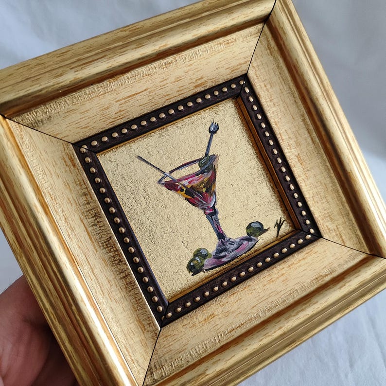 May include: A small painting of a martini glass with olives, framed in a gold frame with a dark brown inner border.