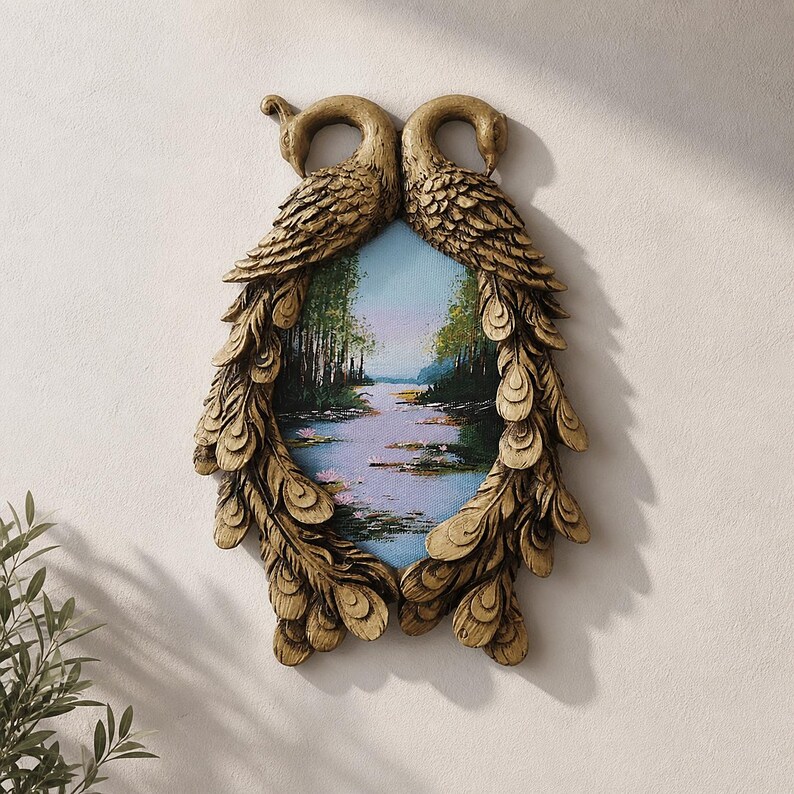 May include: A decorative wall art piece featuring a landscape painting framed by two gold-colored peacock figures. The peacocks' feathers form an oval frame around the painting, which depicts a serene river scene with trees.