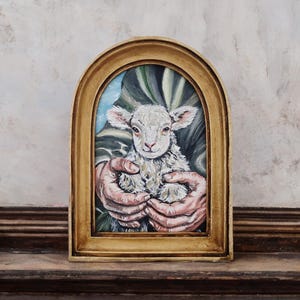 Puede incluir: Pintura enmarcada en un marco dorado en forma de arco, que representa un cordero blanco y esponjoso sostenido en dos manos. El cordero tiene una expresi&oacute;n dulce. El fondo presenta tonos verdes y azules. El marco est&aacute; sobre una superficie marr&oacute;n oscuro.