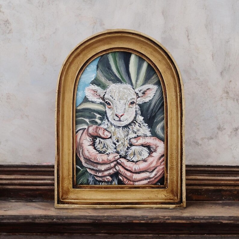 Puede incluir: Pintura enmarcada en un marco dorado en forma de arco, que representa un cordero blanco y esponjoso sostenido en dos manos. El cordero tiene una expresi&oacute;n dulce. El fondo presenta tonos verdes y azules. El marco est&aacute; sobre una superficie marr&oacute;n oscuro.