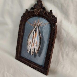Op de afbeelding: Een olieverfschilderij van drie vissen die aan een touwtje hangen, tegen een blauwe achtergrond. Het kunstwerk is ingelijst in een donkerbruine, sierlijke, gewelfde lijst. De vissen hebben zilveren en oranje details.