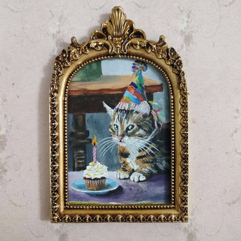 May include: A tabby cat wearing a colorful party hat looks at a cupcake with a lit candle. The cat is sitting on a purple surface in front of a wooden table. The image is framed in a gold ornate frame.