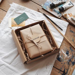 Peut inclure: Une bo&icirc;te en carton marron contenant un cadeau emball&eacute;. Le cadeau est envelopp&eacute; dans du papier brun et ficel&eacute; avec de la ficelle, avec une &eacute;tiquette ronde indiquant "handmade". La bo&icirc;te est sur une surface en bois avec des tubes de peinture, des pinceaux et des cartes de visite.