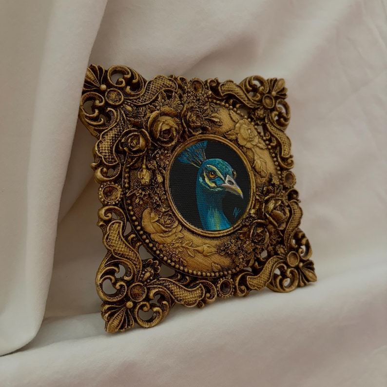 May include: A square, ornate gold-colored frame featuring intricate floral relief patterns surrounding a circular central painting of a peacock head. The peacock is depicted with vibrant blue and green feathers against a dark background.