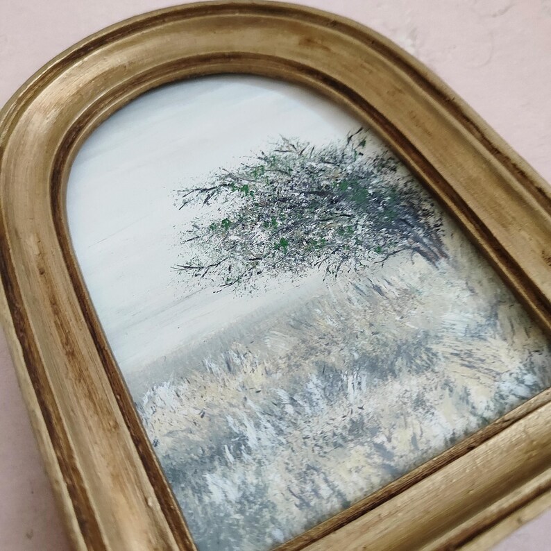 May include: A small, framed landscape painting featuring a tree with green and black foliage against a light background. The painting is set within a gold-colored, arched frame, adding a vintage aesthetic. The artwork depicts a field of tall grass.