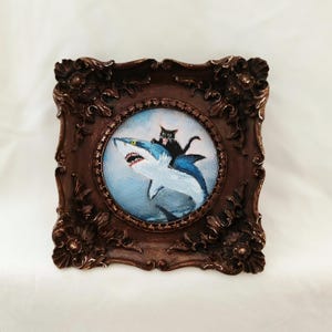 May include: A framed painting depicts a black cat riding a blue shark with an open mouth. The painting is set in a round frame, surrounded by a dark brown, ornate square frame.