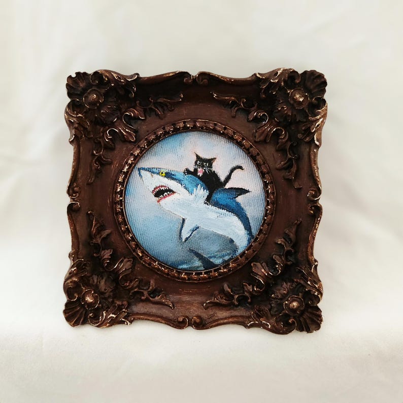 May include: A framed painting depicts a black cat riding a blue shark with an open mouth. The painting is set in a round frame, surrounded by a dark brown, ornate square frame.