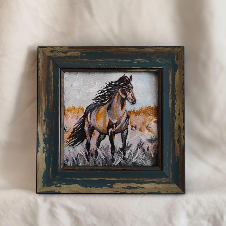 May include: Oil painting of a brown and tan horse with a flowing mane, set against a backdrop of tall grass. The artwork is framed in a distressed, dark teal wooden frame, adding a rustic touch to the piece.