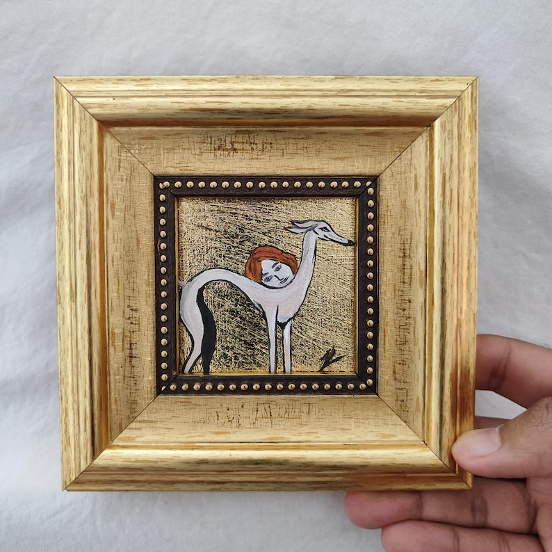 May include: A framed artwork featuring a whimsical illustration of a greyhound-like figure with a human head. The artwork is set against a gold background and framed in a gold-colored wooden frame with a dark brown inner border.