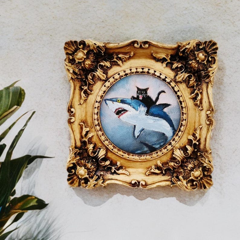 May include: A framed painting featuring a black cat riding a blue and white shark. The artwork is set within a gold-colored, ornate frame. The painting has a whimsical, cartoonish style, perfect for a unique home decor piece.