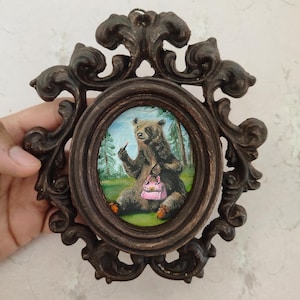 May include: A framed painting of a brown bear wearing a pink purse and holding a lipstick. The bear is sitting in a green grassy area with trees in the background.
