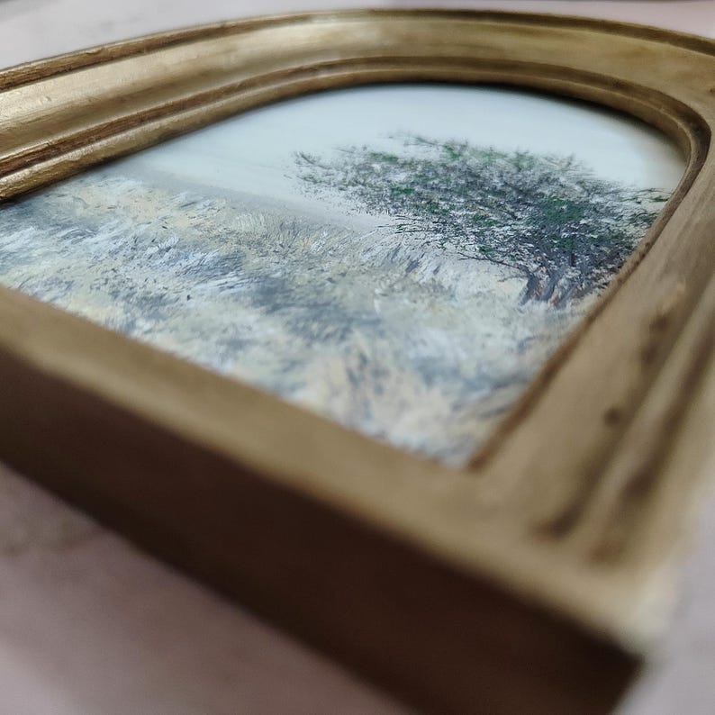 May include: A small, framed landscape painting featuring a tree and field. The painting is set within a gold-colored, arched frame. The artwork depicts a tree with green foliage and a field with muted tones of beige and gray.