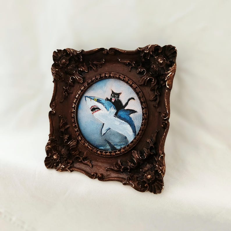 May include: A framed painting depicts a black cat riding a blue and white shark. The artwork is set in a dark brown, ornate, oval frame. The background of the painting is a light blue, creating a whimsical and unique piece of art.