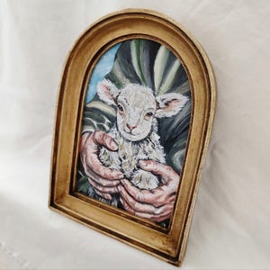 Puede incluir: Pintura enmarcada de un cordero sostenido en manos. El cordero tiene lana blanca y orejas rosas. La pintura est&aacute; en un marco dorado y arqueado. El fondo presenta pinceladas verdes y azules.