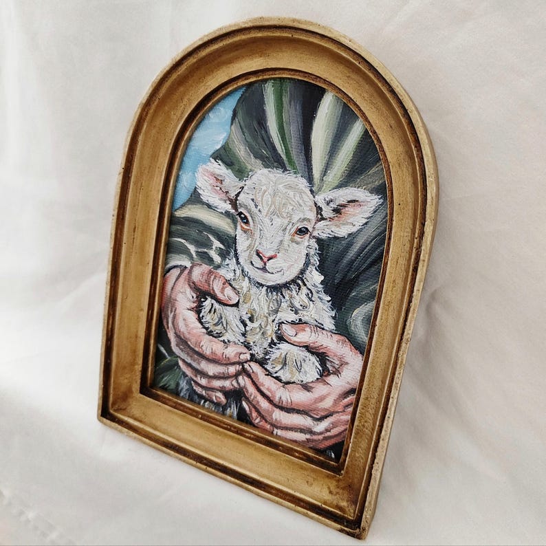 Puede incluir: Pintura enmarcada de un cordero sostenido en manos. El cordero tiene lana blanca y orejas rosas. La pintura est&aacute; en un marco dorado y arqueado. El fondo presenta pinceladas verdes y azules.