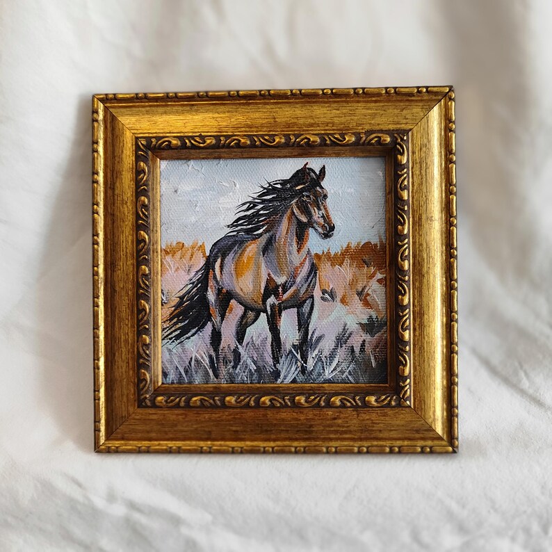 May include: Oil painting in a gold frame depicting a horse in a field. The horse is brown and black with a flowing mane. The background shows tall grass and a light blue sky. The frame has an ornate design.