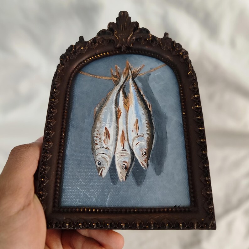 Op de afbeelding: Een ingelijst schilderij toont drie vissen die aan een touwtje hangen tegen een blauwe achtergrond. De vissen zijn geschilderd in zilver, wit en bruin. De lijst is donkerbruin, met een sierlijk, gewelfd ontwerp.