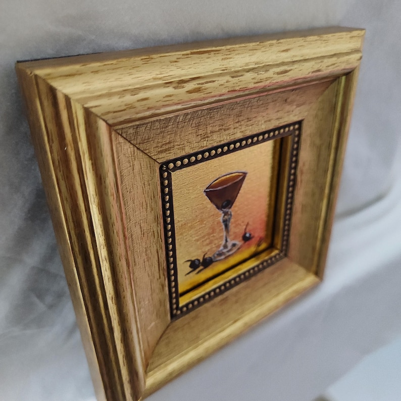 Peut inclure: Peinture encadr&eacute;e d'un verre &agrave; cocktail avec une boisson brun fonc&eacute; et des cerises. L'&oelig;uvre d'art est plac&eacute;e sur un fond dor&eacute; et encadr&eacute;e dans un cadre en bois dor&eacute;.