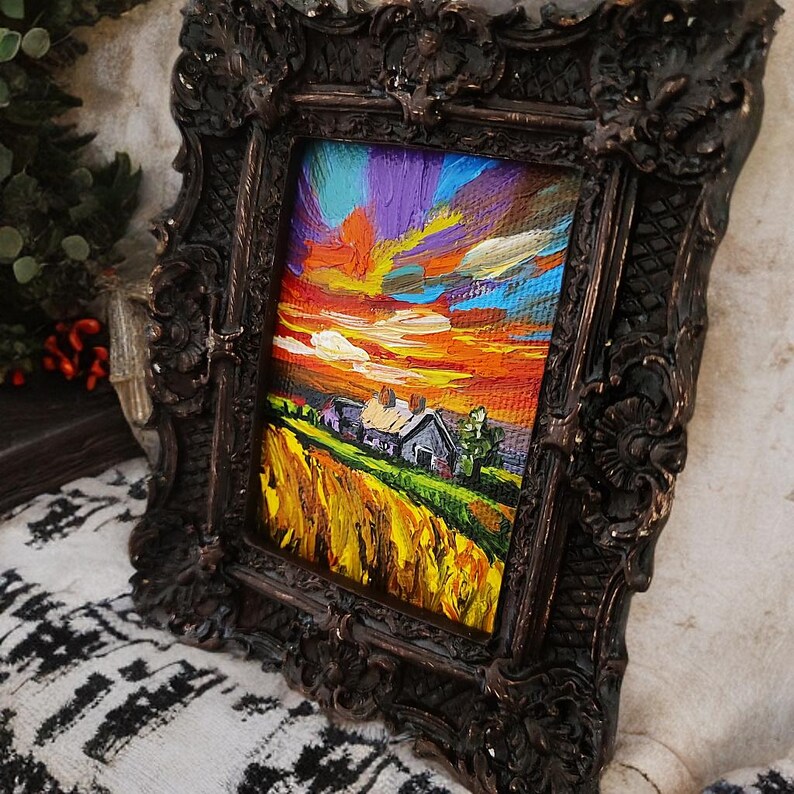 May include: A small, framed landscape painting depicts a vibrant sunset over a rural scene. The artwork features a colorful sky with orange, yellow, and purple hues, and a field of golden wheat. The painting is encased in a dark, ornate frame.