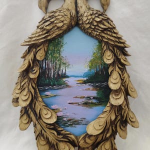May include: A decorative wall hanging featuring a small landscape painting framed by a gold-colored, peacock-feather-shaped border. The painting depicts a river scene with trees and a blue sky. The frame has two bird heads at the top.