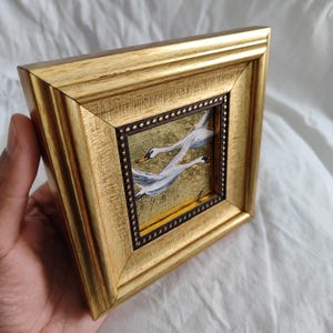 May include: A gold-framed picture with a painting of two white swans flying against a gold background.