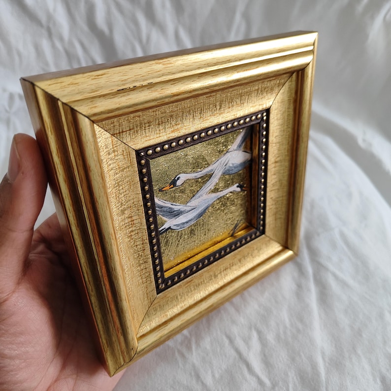 May include: A gold-framed picture with a painting of two white swans flying against a gold background.