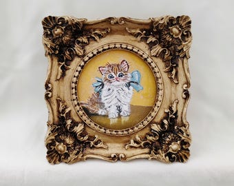 May include: A small, framed painting of a kitten with brown and white fur and blue bow. The painting is set within a decorative, ornate, beige-colored frame. The background of the painting is yellow.