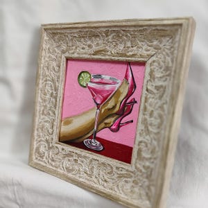 May include: Framed painting of a pink cocktail with a lime wedge, a high-heeled shoe, and a person's arm. The artwork is set against a pink background and framed in a distressed, off-white wooden frame.