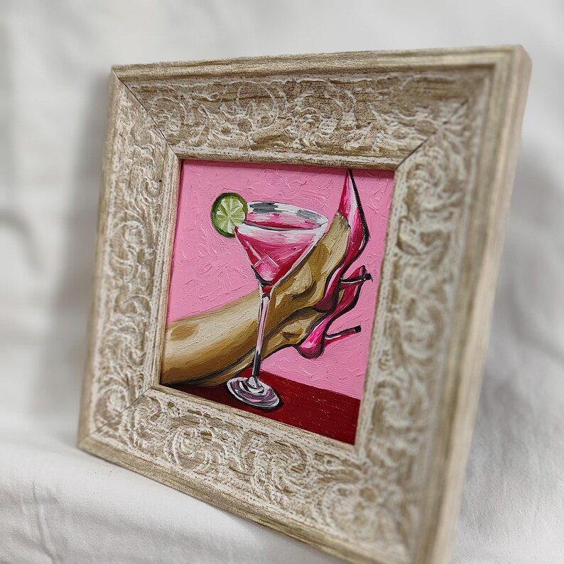 May include: Framed painting of a pink cocktail with a lime wedge, a high-heeled shoe, and a person's arm. The artwork is set against a pink background and framed in a distressed, off-white wooden frame.