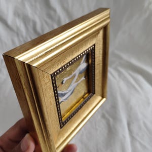 May include: A gold-framed painting of three white swans flying in formation against a gold background. The frame has a decorative border of small gold dots.