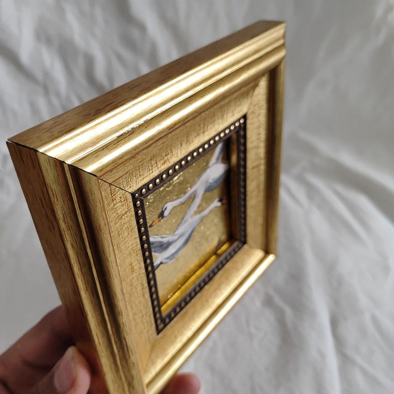 May include: A gold-framed painting of three white swans flying in formation against a gold background. The frame has a decorative border of small gold dots.