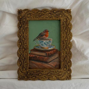 May include: An oil painting in a decorative gold frame featuring a small bird with a red breast perched on a floral teacup and saucer. The teacup rests atop a stack of two brown books against a solid sage green background with visible brushstrokes.