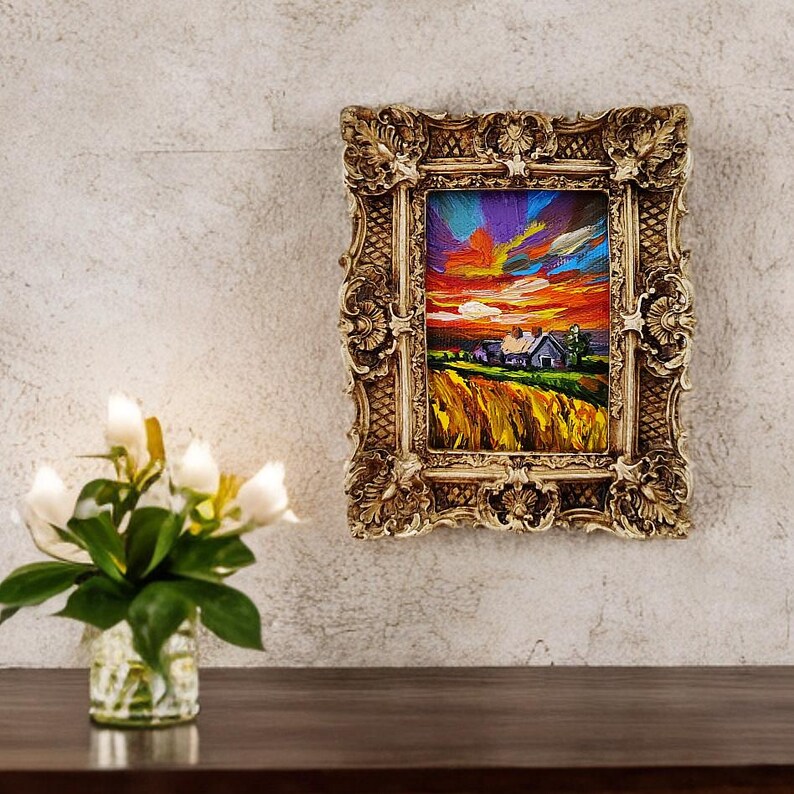 May include: An ornate, gold-colored framed painting depicting a vibrant sunset over a rural landscape. The artwork features a farmhouse, fields of yellow, and a colorful sky with hues of orange, purple, and blue. The frame has detailed scrollwork.