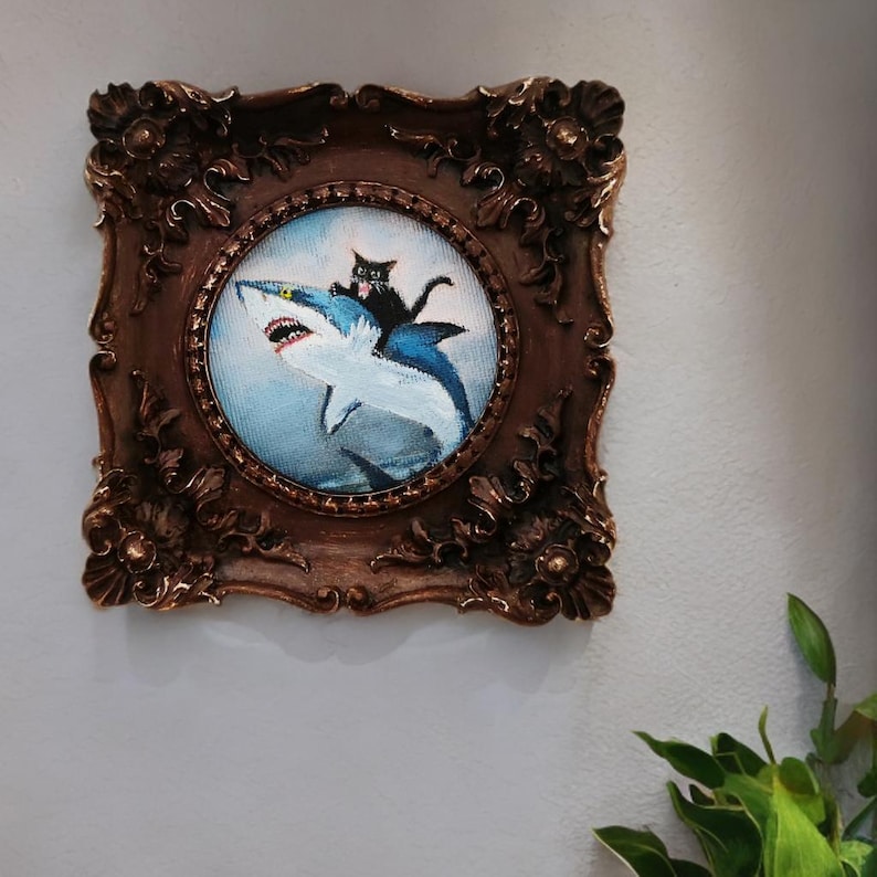 May include: A framed artwork featuring a black cat riding a blue shark. The artwork is in a round frame with a detailed, ornate, brown border. The cat is perched on the shark's back, set against a blue background.
