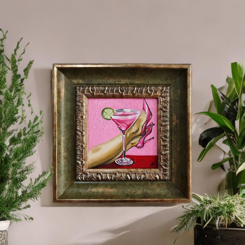 May include: An oil painting in a gold frame depicts a cocktail in a martini glass with a lime wedge, held by a hand wearing a pink high heel. The background is pink and red.