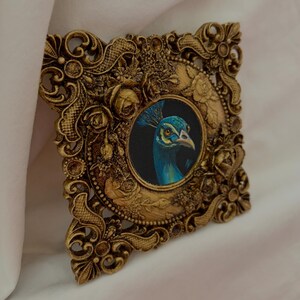 May include: A square wall art piece featuring a central circular painting of a peacock head with blue and green plumage. The artwork is encased in an ornate, textured gold-colored frame decorated with raised floral and scrollwork patterns.