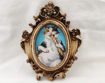 May include: A framed oval artwork featuring a cat with black, white, and tan fur, holding a baguette. The cat is set against a light blue background within a gold-colored ornate frame. The frame has decorative floral and scroll details.