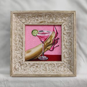 May include: Framed painting of a pink cocktail with a lime garnish, a high-heeled shoe, and a hand. The artwork is set against a pink background with a red base, framed in a distressed, ornate, light-colored wood frame.