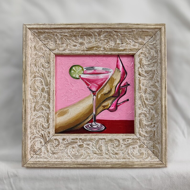 May include: Framed painting of a pink cocktail with a lime garnish, a high-heeled shoe, and a hand. The artwork is set against a pink background with a red base, framed in a distressed, ornate, light-colored wood frame.