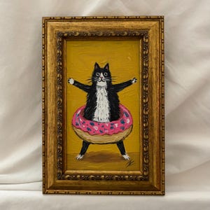 May include: A framed oil painting featuring a black and white cat with wide eyes standing inside a large pink donut with colorful sprinkles. The cat has its arms outstretched against a solid mustard yellow background, encased in an ornate gold frame.